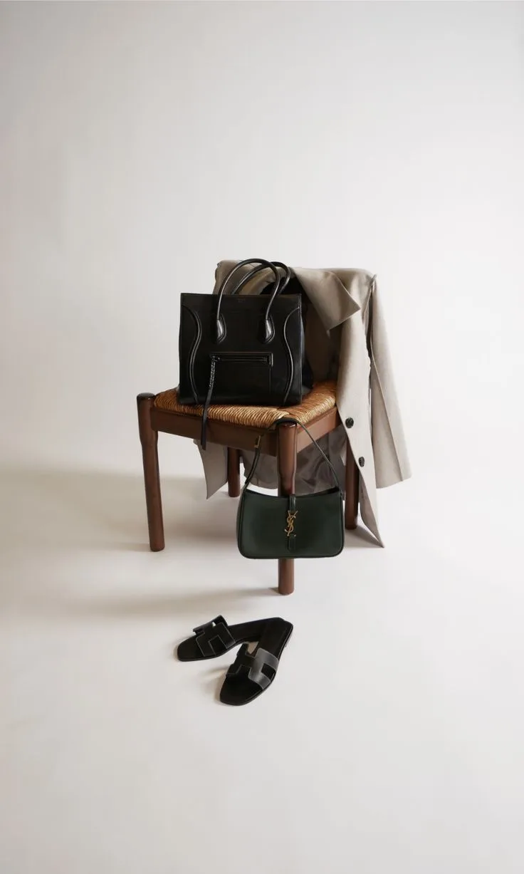 A black leather tote, a linen blazer, a green shoulder bag and leather sandals arranged on a wooden chair